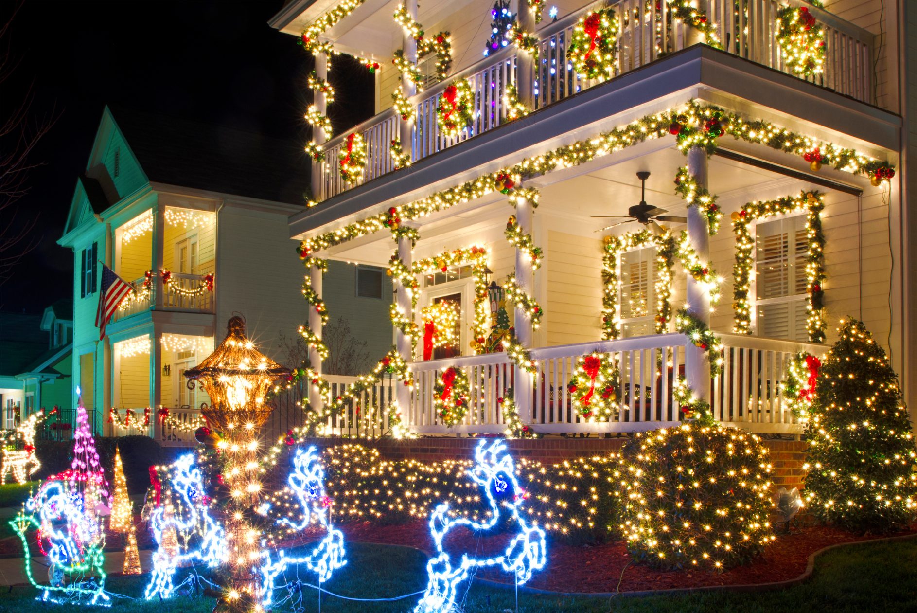 Home in neighborhood decorated with garlands and bright white and colorful LED Christmas lights, including a porch and light-up reindeer figures on the lawn.