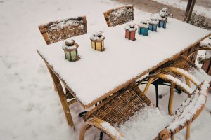 An outdoor cafe table and wicker chairs completely covered in a fresh layer of white snow. Five colorful vintage-style glass lanterns sit on the tabletop, partially buried in the snow, creating a quiet and cold winter scene.